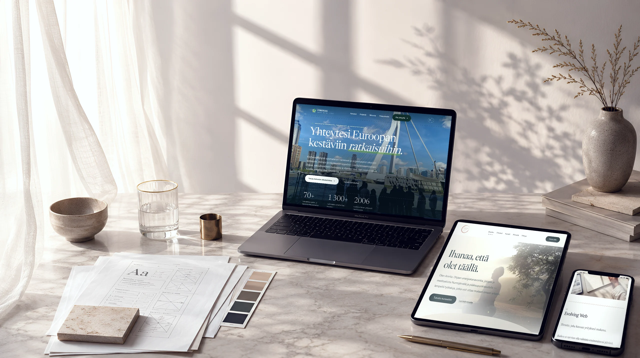 Kannettava tietokone, tabletti ja puhelin marmoripöydällä. Näytöillä on Studio Titan suunnittelemia sivustoja: FinDera Consulting, Amrita Gian ja Studio Titan Evolving Web -palvelu. Pöydällä on myös luonnoksia, värimalleja ja keramiikkakulho, ja seinällä näkyy lehtien varjo.